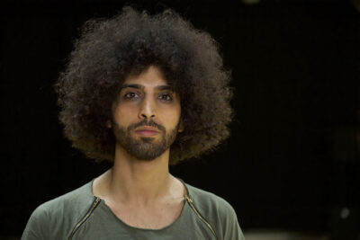 Das Bild zeigt den Tänzer und Choreografen Akiles, der in Berlin lebt und ursprünglich aus dem Irak kommt. Er blickt auf dem Portraitbild direkt in die Kamera.