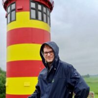 Das Bild zeigt den Musiker Calvin Lanz. Im Bild steht er vor einem gelb-rot gestreiften Turmhaus - ein Leuchtturm? Er trägt einen dunkelblauen Regenmantel und hat die Kapuze aufgesetzt.
