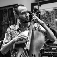 Das Foto zeigt den Cellisten Gabor Hartyani. Er spielt gerade Cello. Das Foto ist in schwarz/weiß. Im Hintergrund hängen große gemalte und gerahmte Bilder an den Wänden.