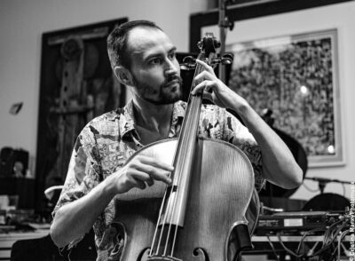 Das Foto zeigt den Cellisten Gabor Hartyani. Er spielt gerade Cello. Das Foto ist in schwarz/weiß. Im Hintergrund hängen große gemalte und gerahmte Bilder an den Wänden.