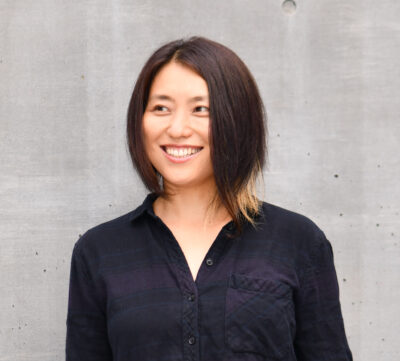 Das Bild zeigt ein Portrait der japanischen Komponistin Reiko Yamada. Sie trägt ein schwarzes Oberteil und lächelt.