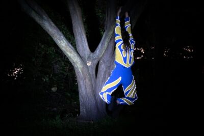 eine Person in blau-gelbem Kostüm hält sich hängend an einem Baum fest. Es ist dunkel.